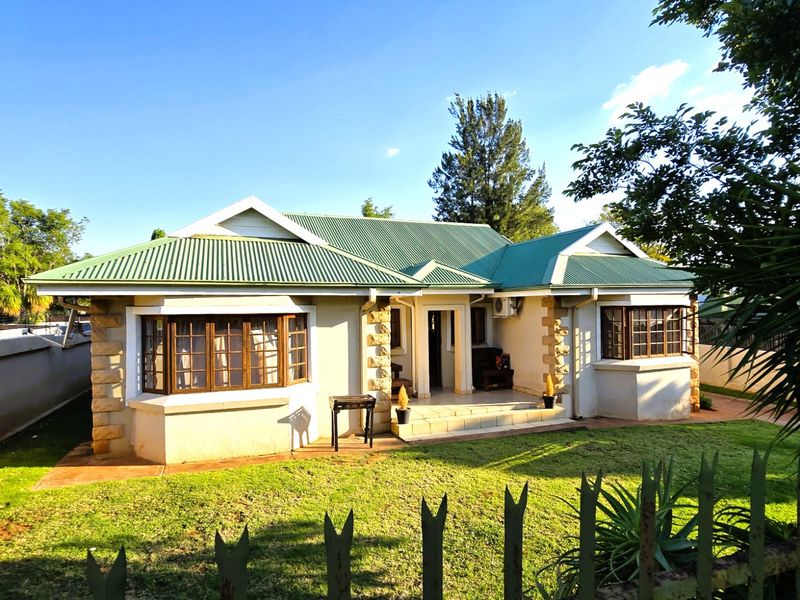 A charming single-story house featuring a green corrugated metal roof, stone corner detailing, and classic bay windows, set within a well-maintained garden.