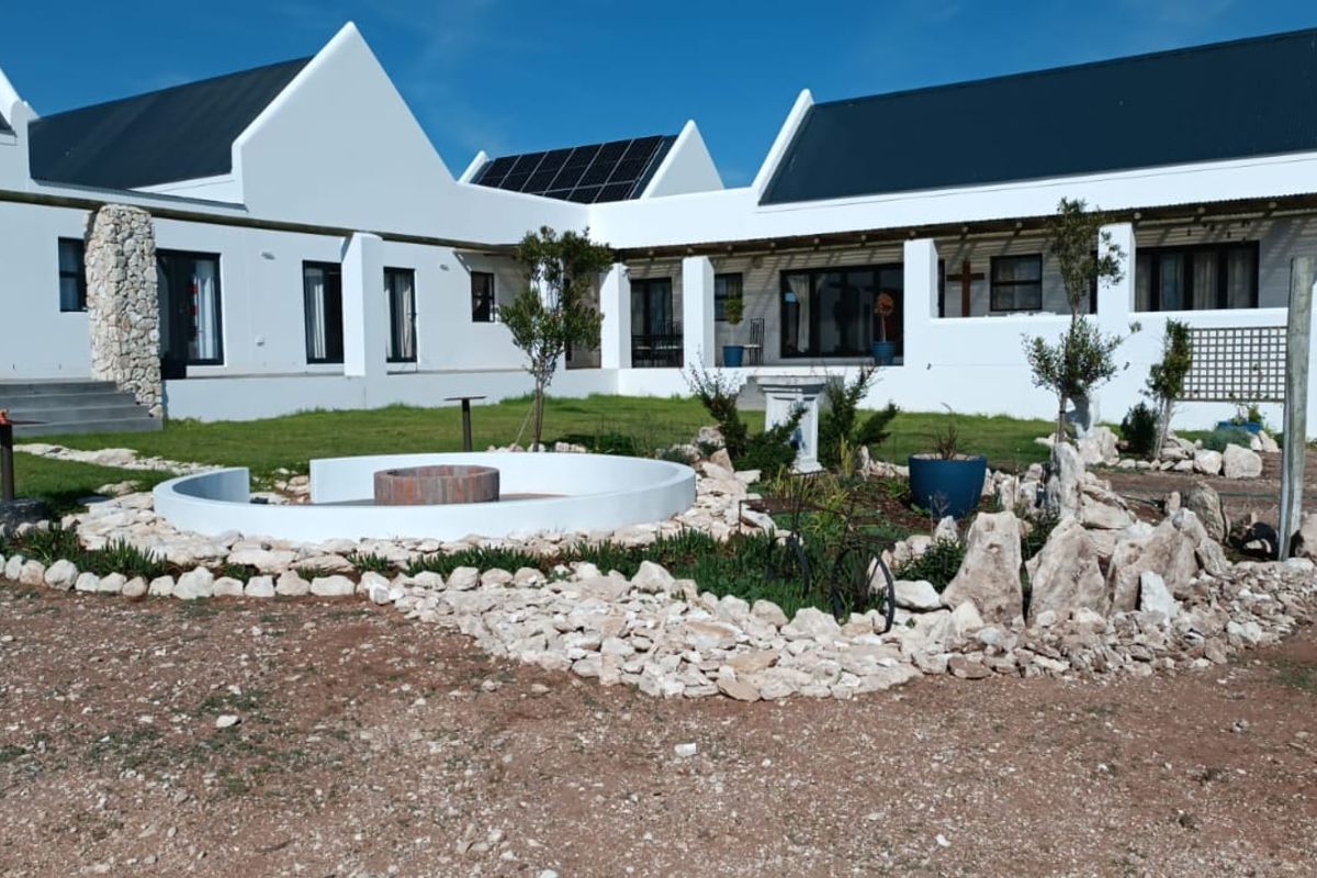 A well-maintained rear exterior view of a modern house featuring a white facade, metal roofing, solar panels, and a landscaped garden with a fire pit.
