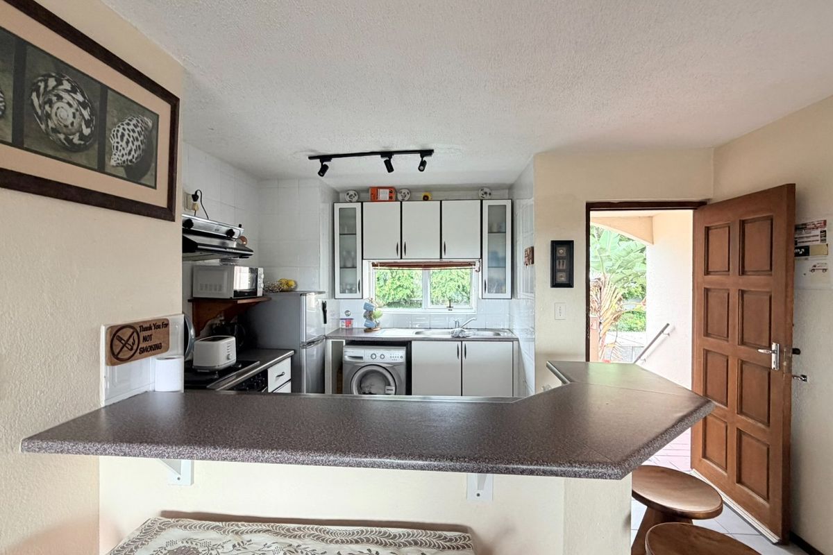 Compact kitchen featuring white cabinetry, laminate countertops, tiled backsplash, and integrated laundry plumbing, with direct access to an outdoor area.