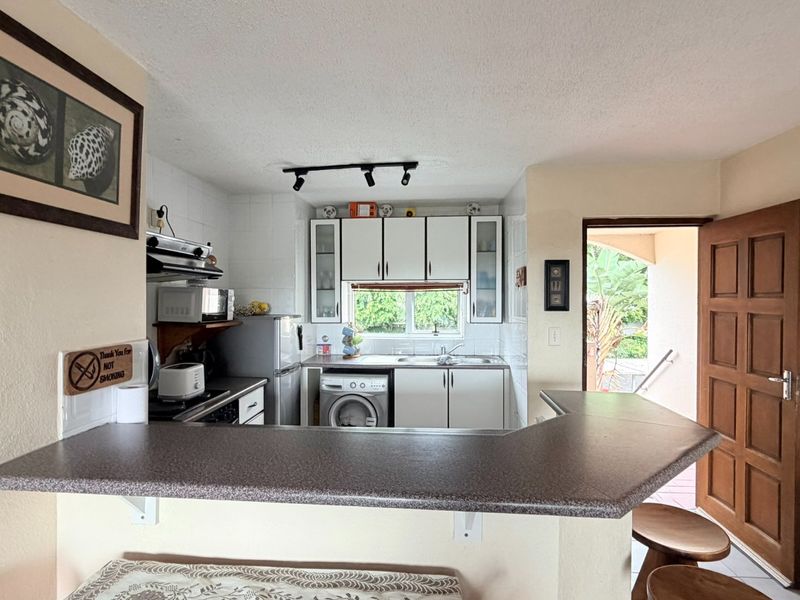 Compact kitchen featuring white cabinetry, laminate countertops, tiled backsplash, and integrated laundry plumbing, with direct access to an outdoor area.