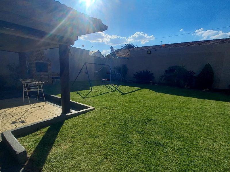 A spacious rear garden featuring a well-maintained lawn, a covered patio area, and a built-in braai, perfect for outdoor entertaining.