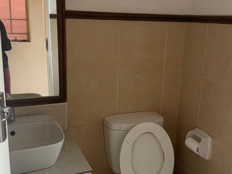 A functional bathroom featuring a vessel sink with vanity cabinet, wall-mounted mirror, and standard toilet, finished with neutral ceramic wall and floor tiling.