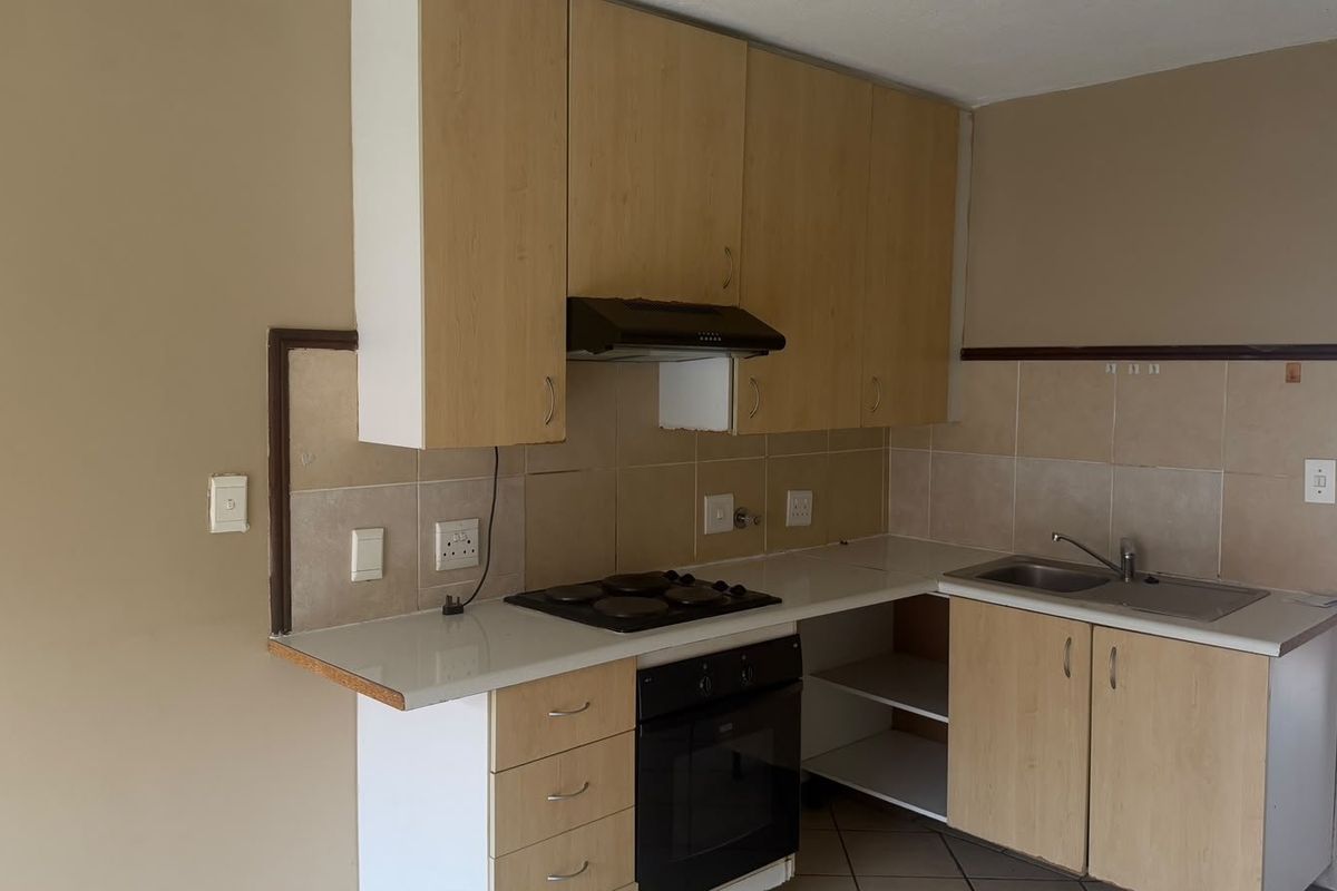 A compact kitchen featuring light wood-finish cabinetry, a built-in oven with electric hob, extractor fan, and a stainless steel sink with tiled backsplash.