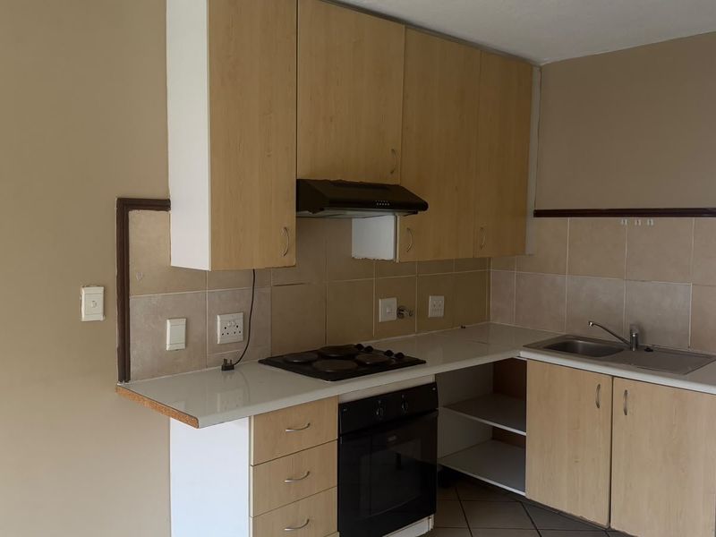 A compact kitchen featuring light wood-finish cabinetry, a built-in oven with electric hob, extractor fan, and a stainless steel sink with tiled backsplash.