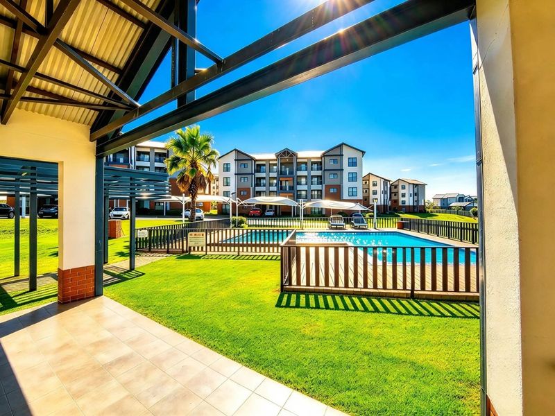 A well-maintained communal pool area featuring a fenced swimming pool, manicured lawn, and a covered tiled patio area within an apartment complex.