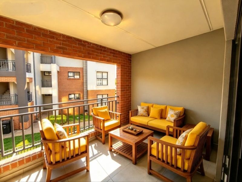 A covered balcony featuring exposed brick accents, tiled flooring, and a metal balustrade, overlooking the apartment complex grounds.