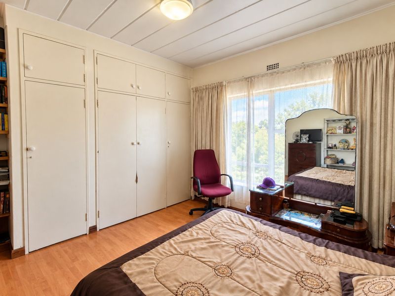 A well-lit bedroom featuring extensive built-in cupboard storage, laminate flooring, and a large window with curtain treatments.