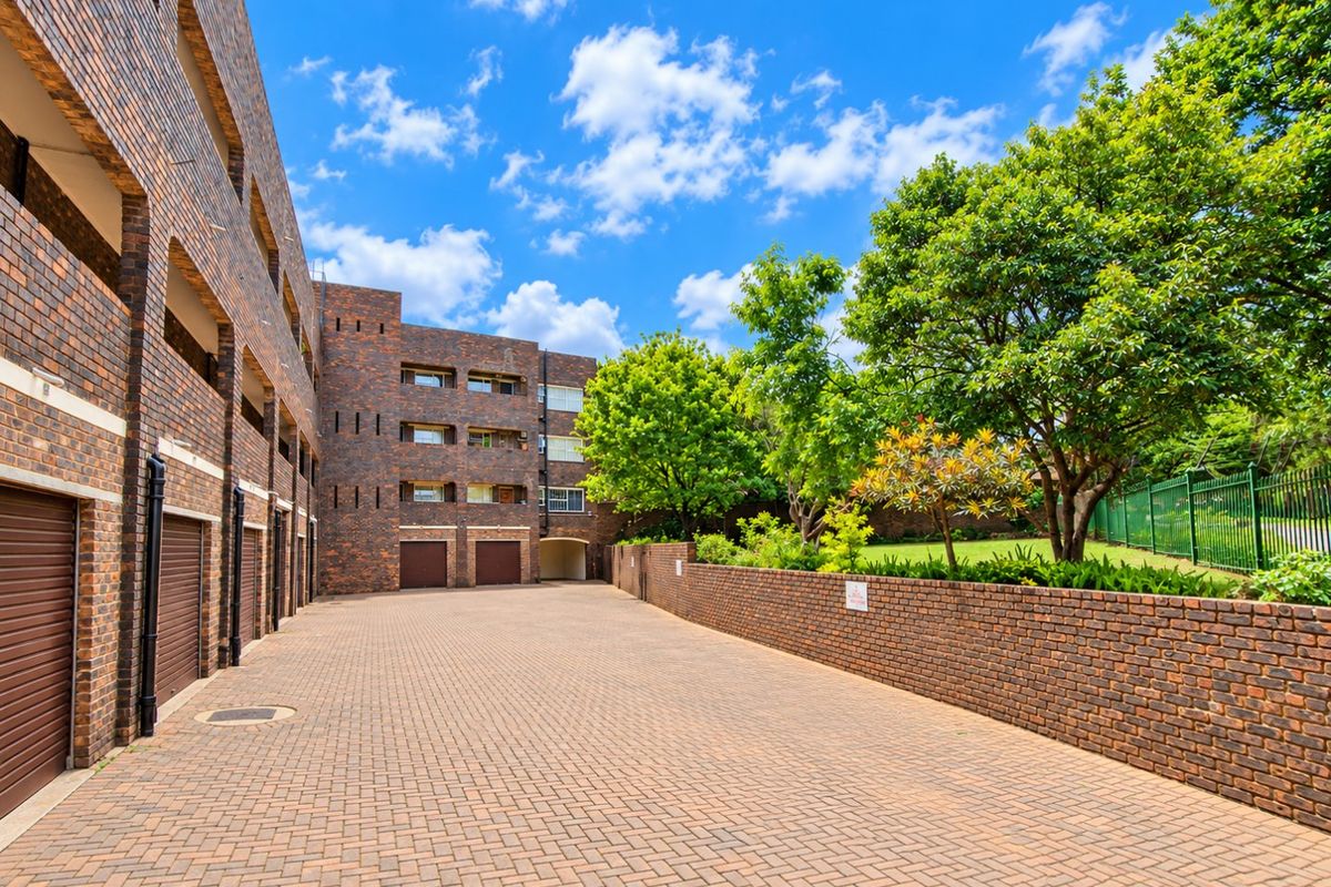 A well-maintained exterior rear view of the apartment complex featuring brick paving, face brick building facades, integrated garage access, and a landscaped garden area.