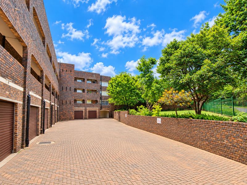 A well-maintained exterior rear view of the apartment complex featuring brick paving, face brick building facades, integrated garage access, and a landscaped garden area.