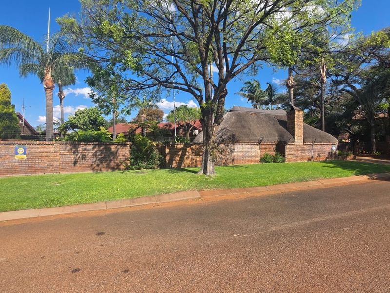 Street view of the property featuring a brick boundary wall, established trees, and a traditional thatch roof structure.