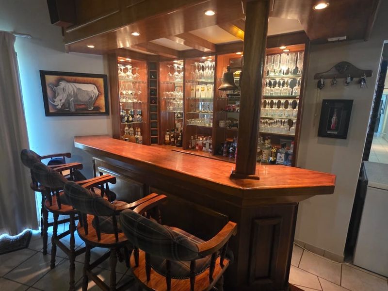 A dedicated home bar area featuring a custom-built wooden counter, integrated glass shelving with display lighting, and durable tiled flooring.