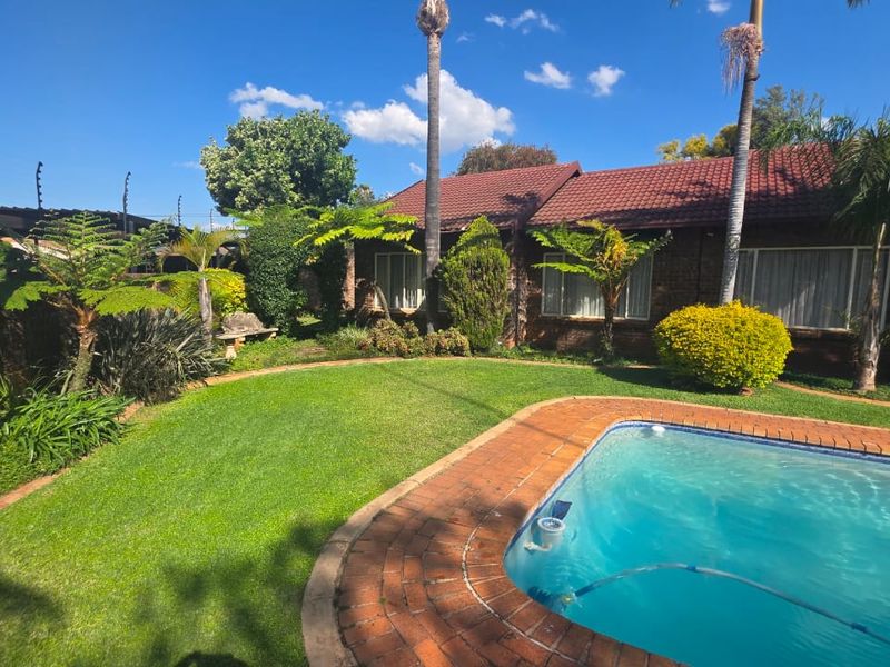 A well-maintained backyard featuring a swimming pool with brick coping, surrounded by a lush lawn and established garden beds against a brick house.