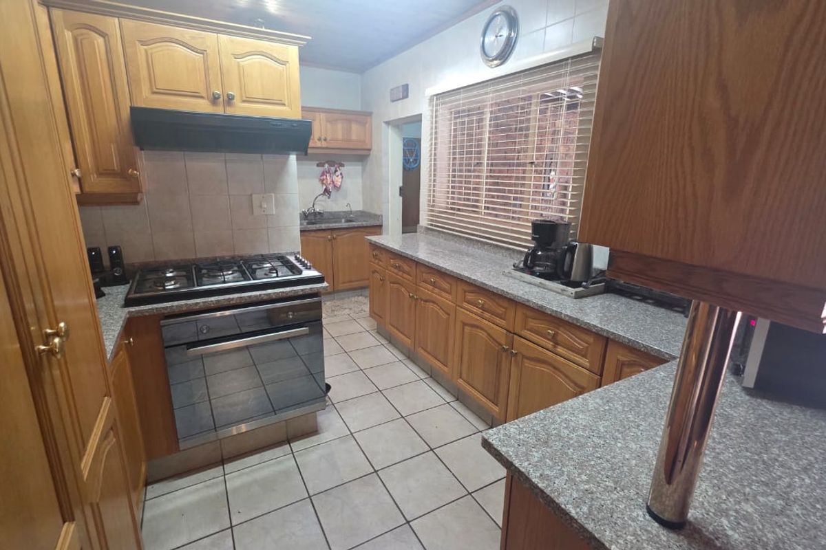 A functional kitchen featuring wooden cabinetry, granite countertops, a gas hob, built-in oven, and tiled flooring with a separate scullery area visible.