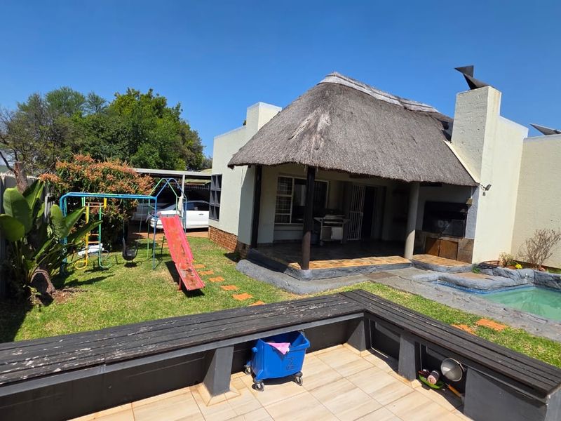 Rear exterior view featuring a thatched roof patio, built-in braai area, swimming pool, and a paved seating area with a built-in bench.