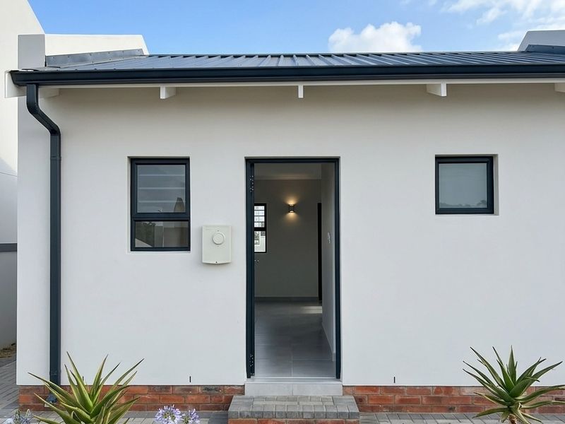 A clean, modern rear exterior view featuring a paved walkway, brick plinth detailing, and dark aluminium window frames against a crisp white facade.