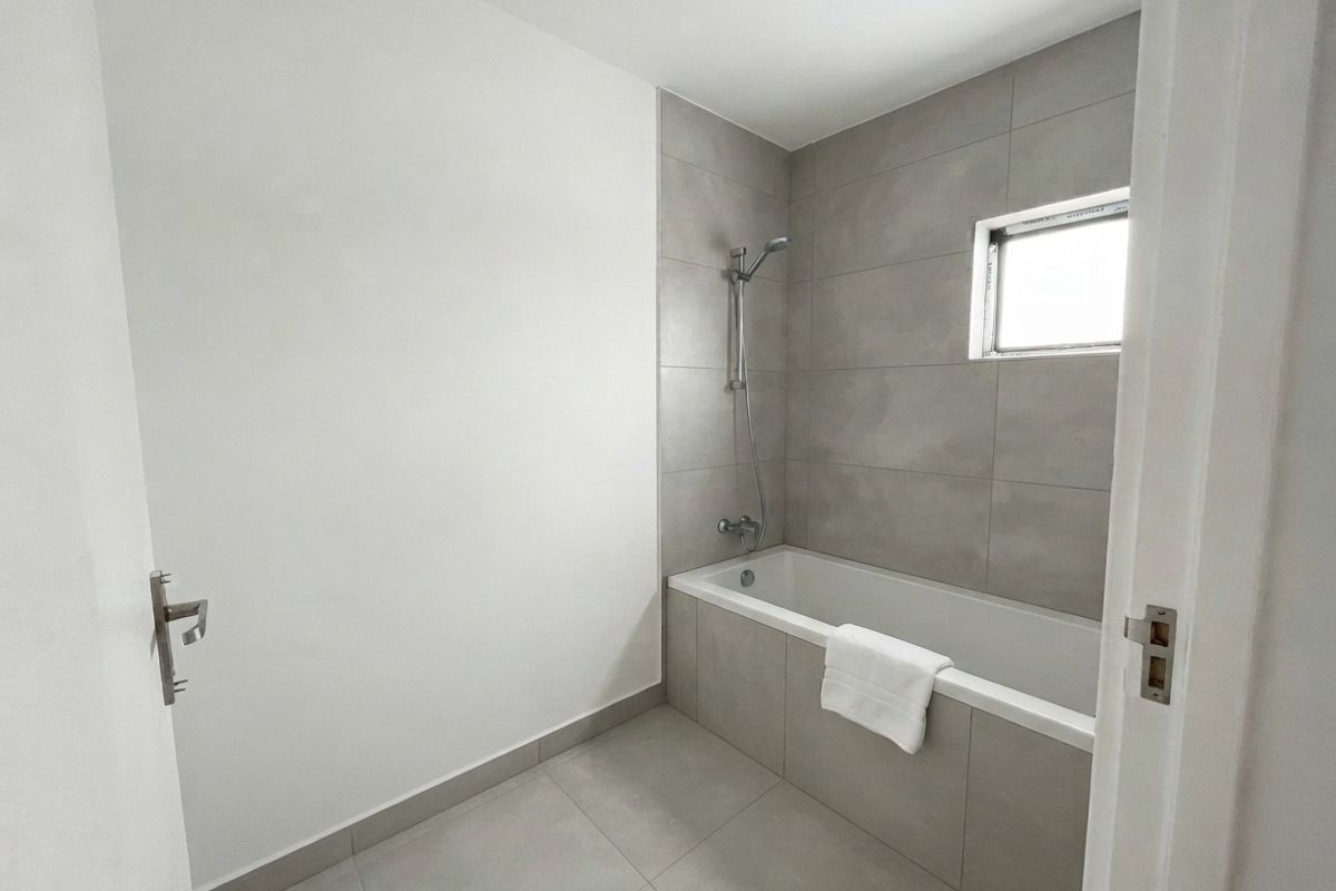 A clean, modern bathroom featuring neutral grey floor and wall tiles, a built-in bathtub with a handheld shower fixture, and a small window.