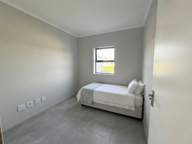 A compact, neutral-toned bedroom featuring modern tiled flooring, clean finishes, and a single window providing natural light.