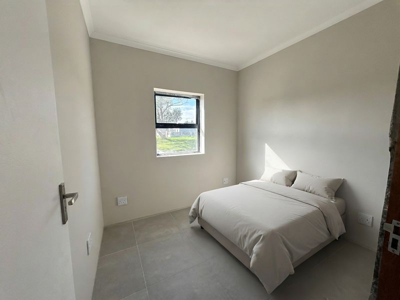 A clean, neutral bedroom featuring large floor tiles, modern aluminium window frames, and minimalist finishes, offering a bright and functional living space.