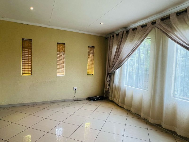 A bright, empty living room featuring neutral tiled flooring, recessed downlighting, and large windows fitted with curtains and vertical blinds.