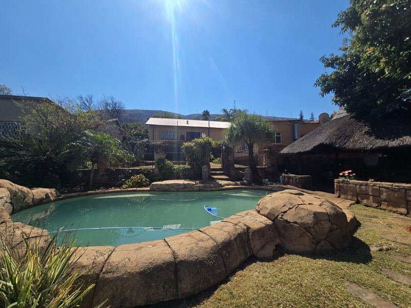 Outdoor pool area featuring a rock-style swimming pool, stone retaining walls, and a traditional thatched lapa, set within a landscaped garden.