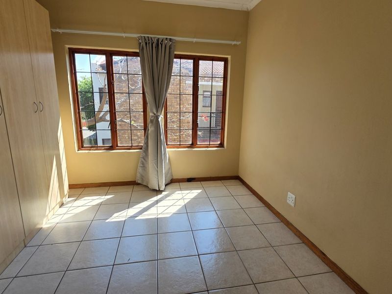 A bright, empty bedroom featuring tiled flooring, built-in cupboards, and large wooden-framed windows allowing for natural light.