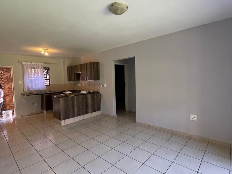 An open-plan living and kitchen area featuring neutral tiled flooring, built-in kitchen cabinetry with a breakfast bar, and a clean, minimalist layout.