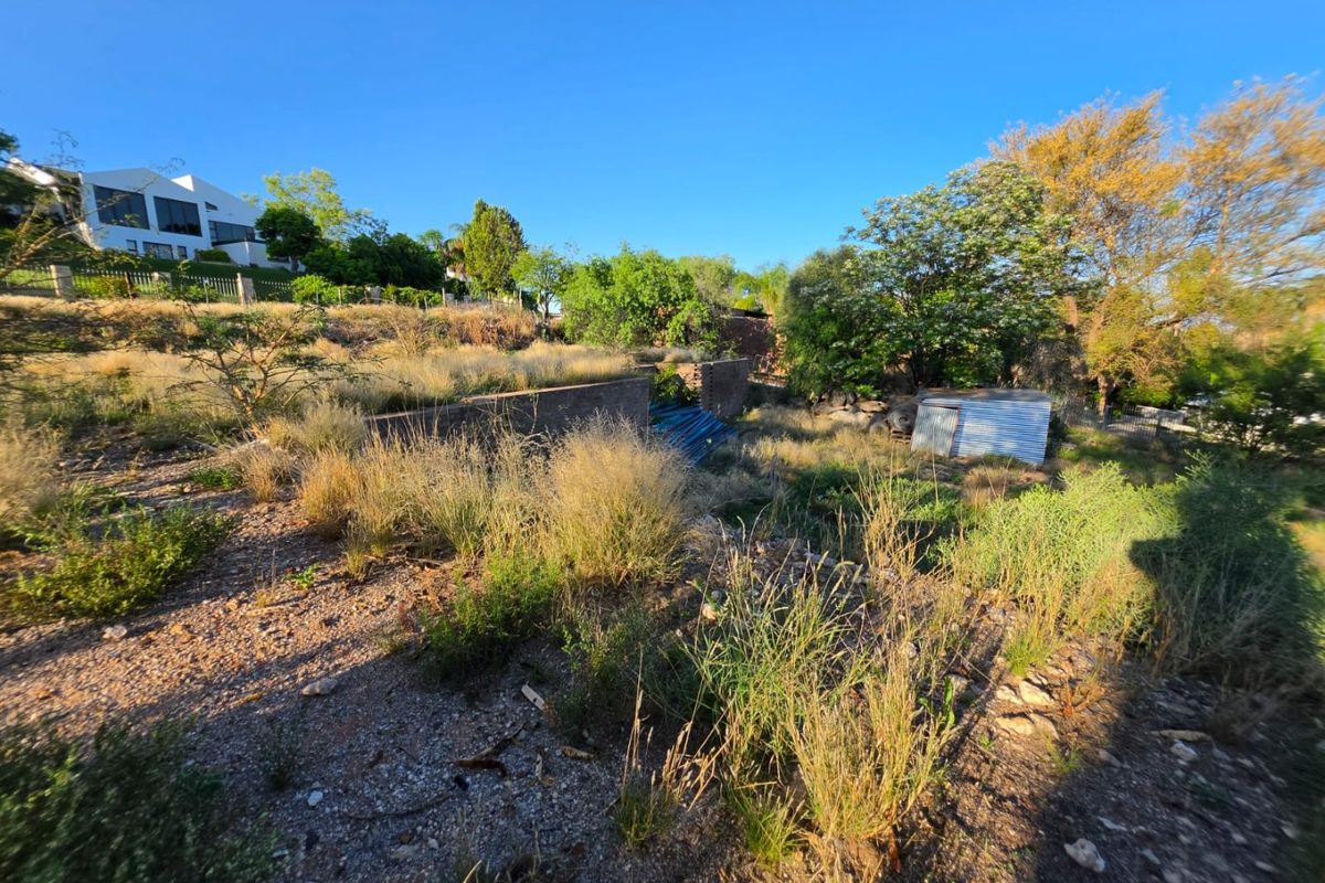 A view of a vacant residential plot featuring a retaining wall, overgrown vegetation, and a small corrugated metal storage structure.