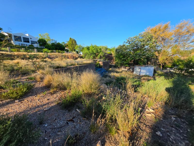 A view of a vacant residential plot featuring a retaining wall, overgrown vegetation, and a small corrugated metal storage structure.