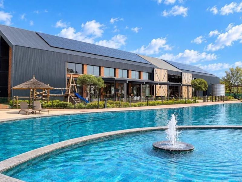 A well-maintained outdoor pool area featuring a central water fountain, paved decking, and a modern building with solar panels and brick accents.