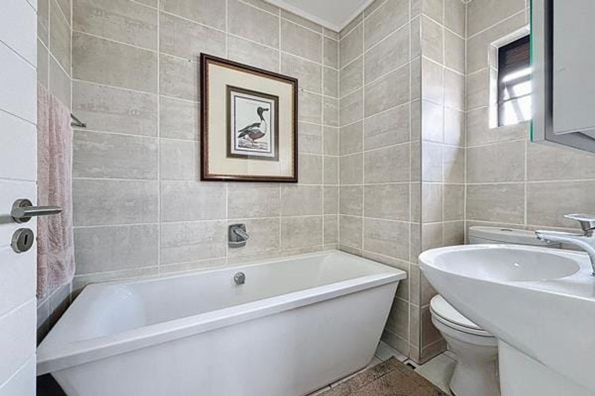A clean, well-maintained bathroom featuring neutral ceramic wall tiles, a built-in bathtub, a pedestal washbasin, and a standard toilet.