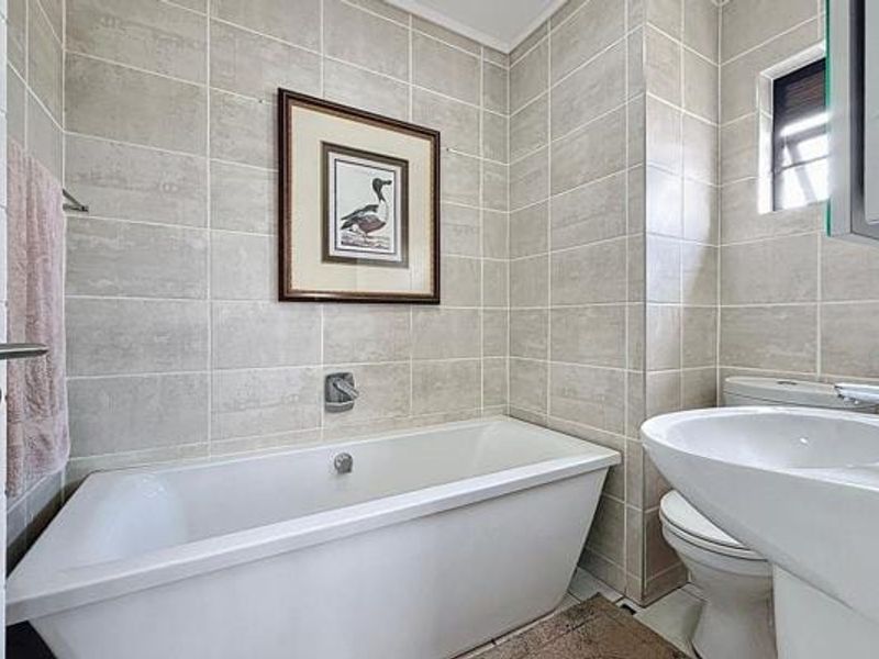 A clean, well-maintained bathroom featuring neutral ceramic wall tiles, a built-in bathtub, a pedestal washbasin, and a standard toilet.