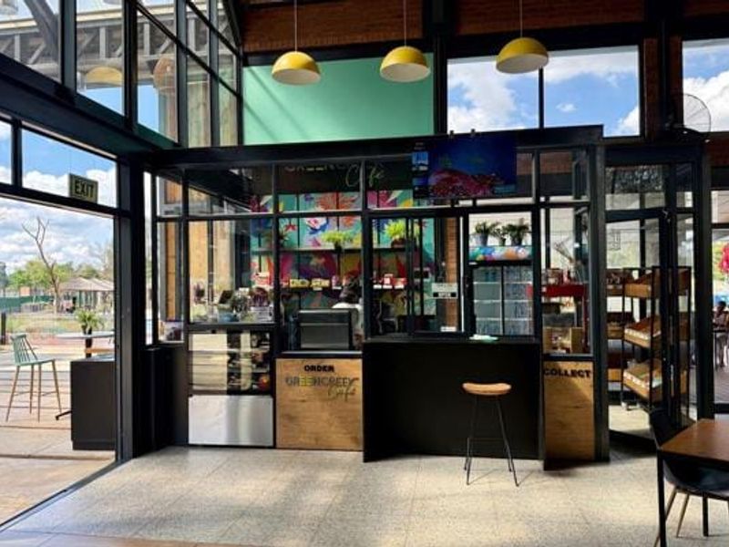 A modern commercial cafe space featuring industrial-style glass partitions, metal framing, tiled flooring, and integrated service counters with pendant lighting.