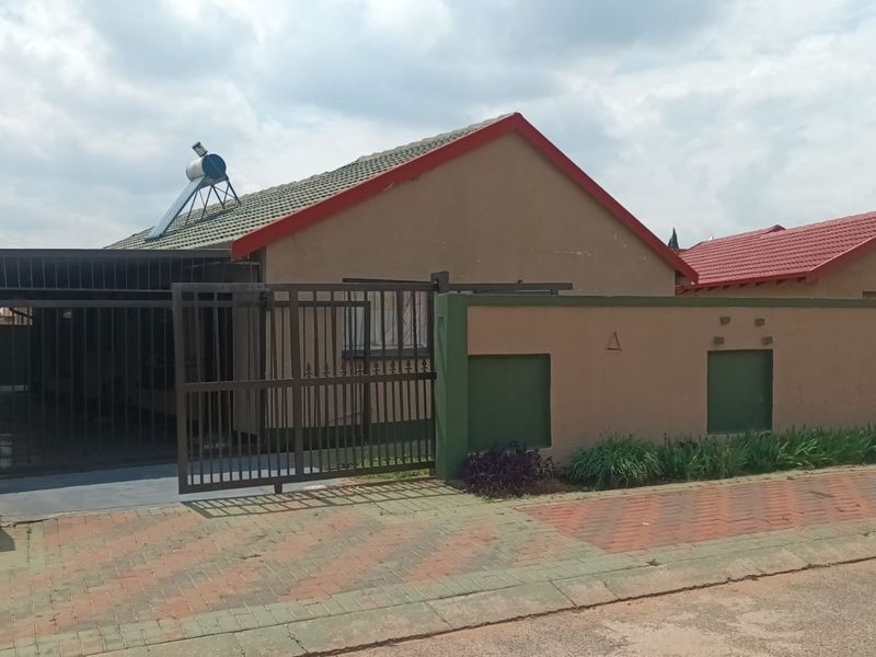 Front exterior view of a single-story house featuring a tiled roof, solar water heater, paved driveway, and a secure metal gate with perimeter walling.