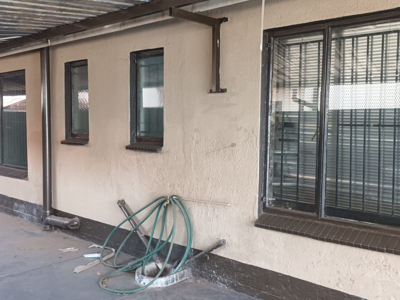 A covered outdoor patio area featuring concrete flooring, security-barred windows, and an external water tap, showing signs of wear on the exterior wall finish.
