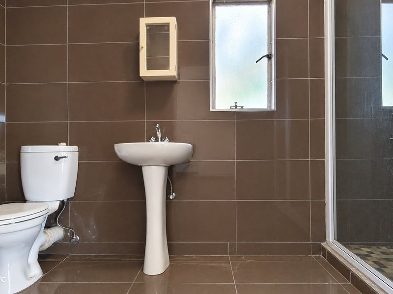 A functional bathroom featuring brown wall and floor tiles, a pedestal basin, a toilet, and a glass-enclosed shower.