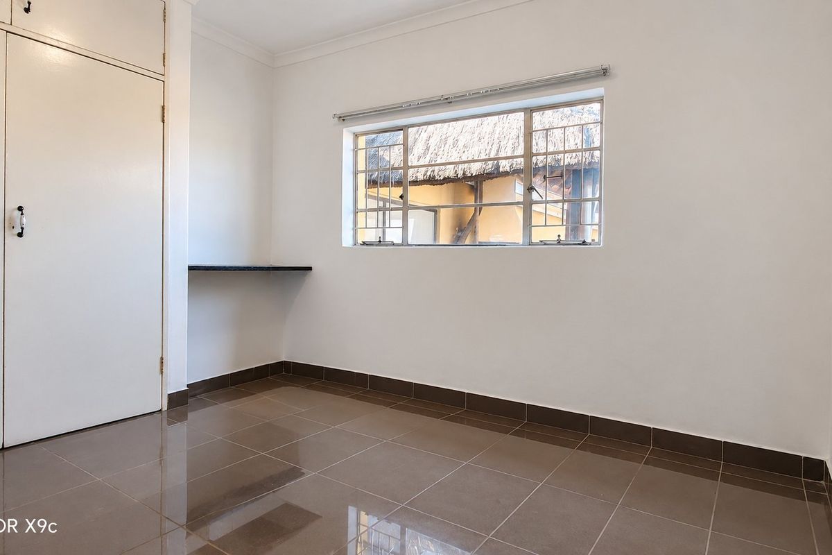 A clean, empty bedroom featuring tiled flooring, built-in cupboards, and a built-in desk area with a window overlooking the property exterior.