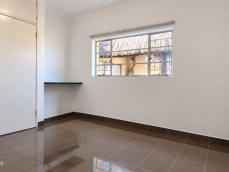 A clean, empty bedroom featuring tiled flooring, built-in cupboards, and a built-in desk area with a window overlooking the property exterior.