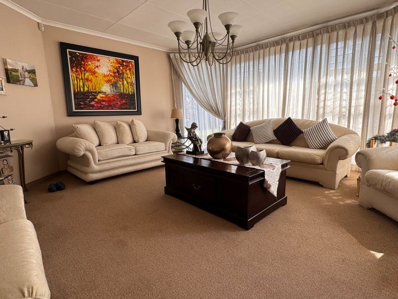 A spacious living room featuring neutral carpeting, a central chandelier, and large windows draped with floor-to-ceiling curtains.