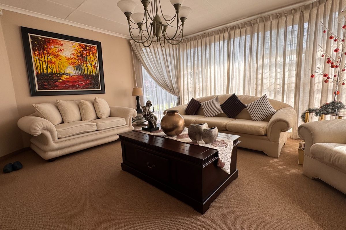A comfortable living room featuring neutral carpeting, a classic chandelier, and large windows dressed with floor-to-ceiling curtains.
