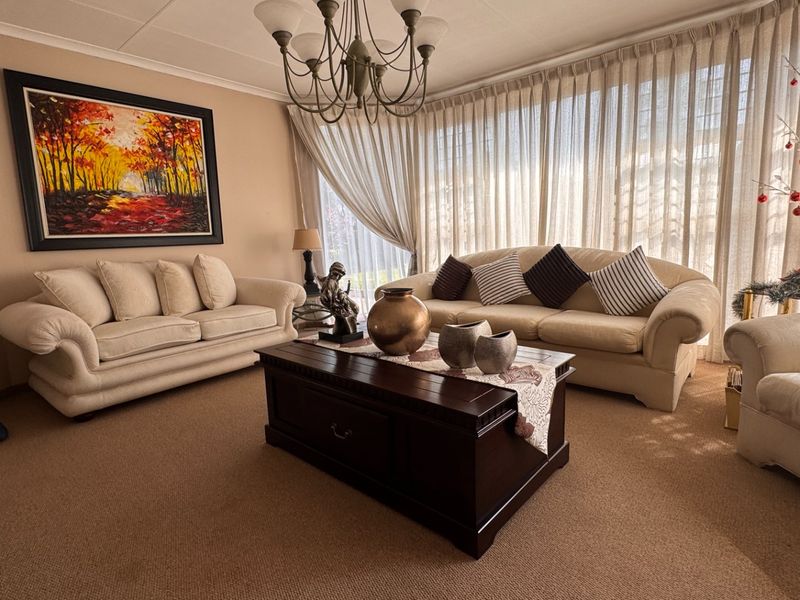A comfortable living room featuring neutral carpeting, a classic chandelier, and large windows dressed with floor-to-ceiling curtains.
