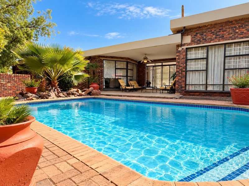 A well-maintained outdoor pool area featuring brick paving, a covered patio with a ceiling fan, and a classic brick exterior facade.