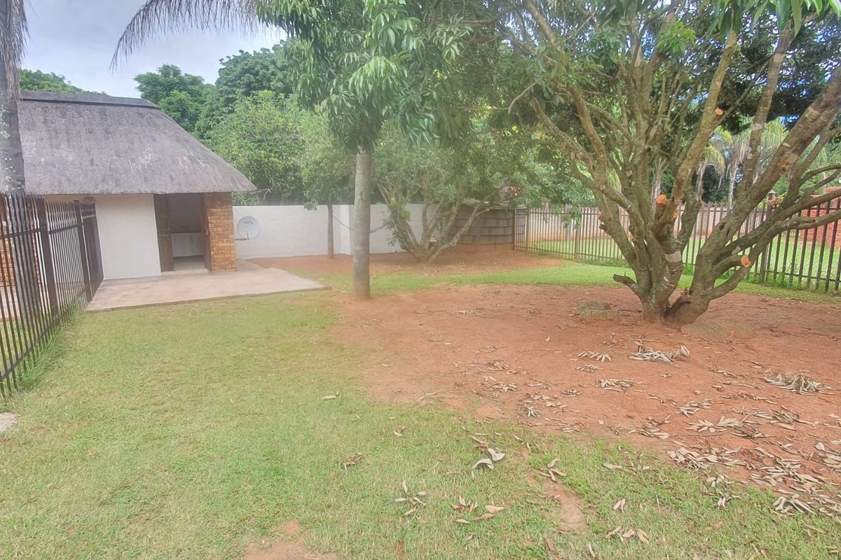 A rear garden area featuring a thatch-roofed structure, concrete patio, and metal perimeter fencing, with established trees and open lawn space.