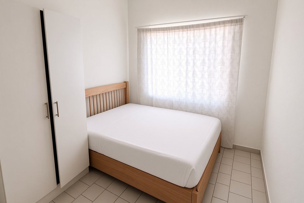 A compact bedroom featuring tiled flooring, a built-in wardrobe, and a window with sheer curtains, offering a clean and functional living space.