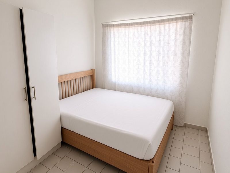 A compact bedroom featuring tiled flooring, a built-in wardrobe, and a window with sheer curtains, offering a clean and functional living space.