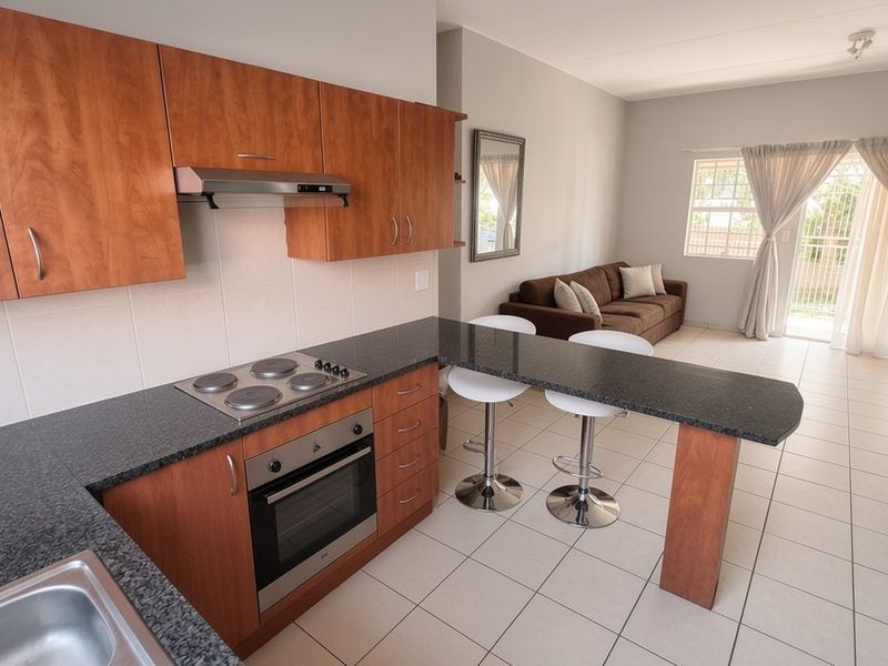 Open-plan kitchen and living area featuring wood-finish cabinetry, granite countertops, built-in appliances, and tiled flooring leading to a balcony.
