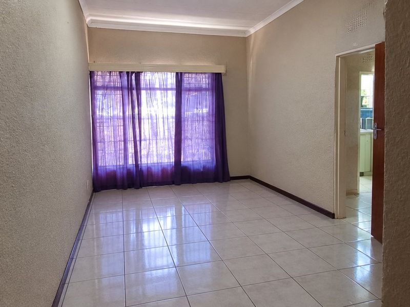 An empty living room featuring neutral tiled flooring, cove cornicing, and a large window with security bars.