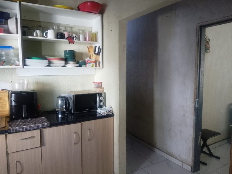 A compact kitchen area featuring built-in cabinetry, granite countertops, and open shelving, leading into an adjacent hallway with tiled flooring.