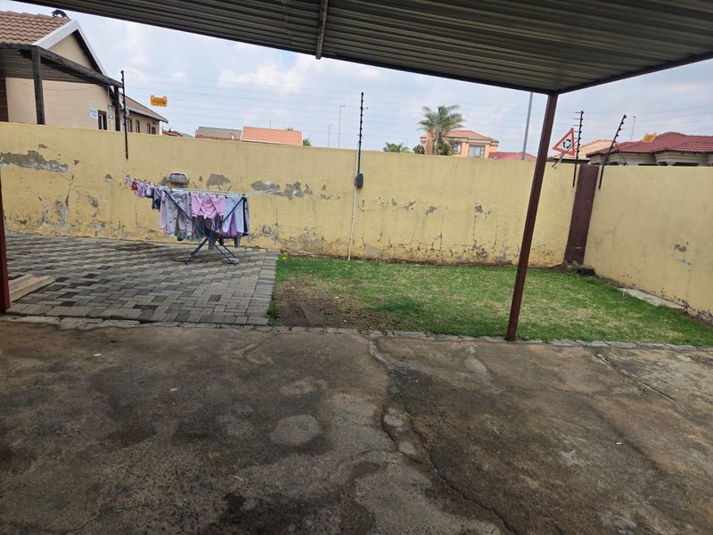 A rear outdoor area featuring a covered patio, paved section, small lawn, and perimeter wall with electric fencing.