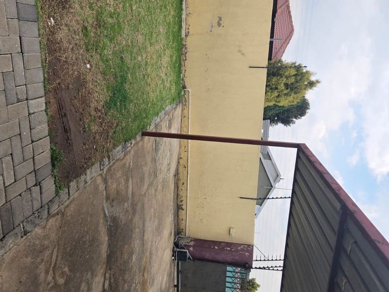 A rear outdoor area featuring a paved driveway, a small lawn, a carport structure, and a perimeter wall with electric fencing.