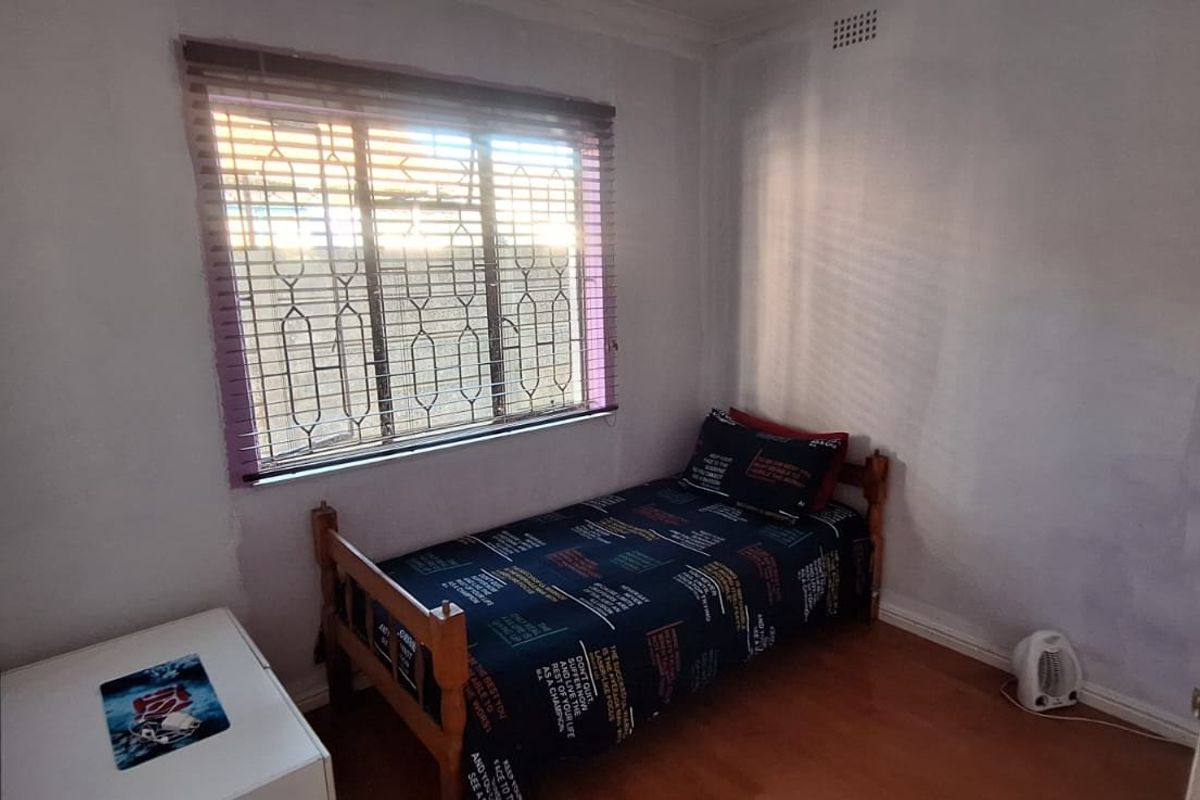 A compact bedroom featuring laminate flooring, a window with security bars, and venetian blinds. The room requires cosmetic updates to the wall finishes.